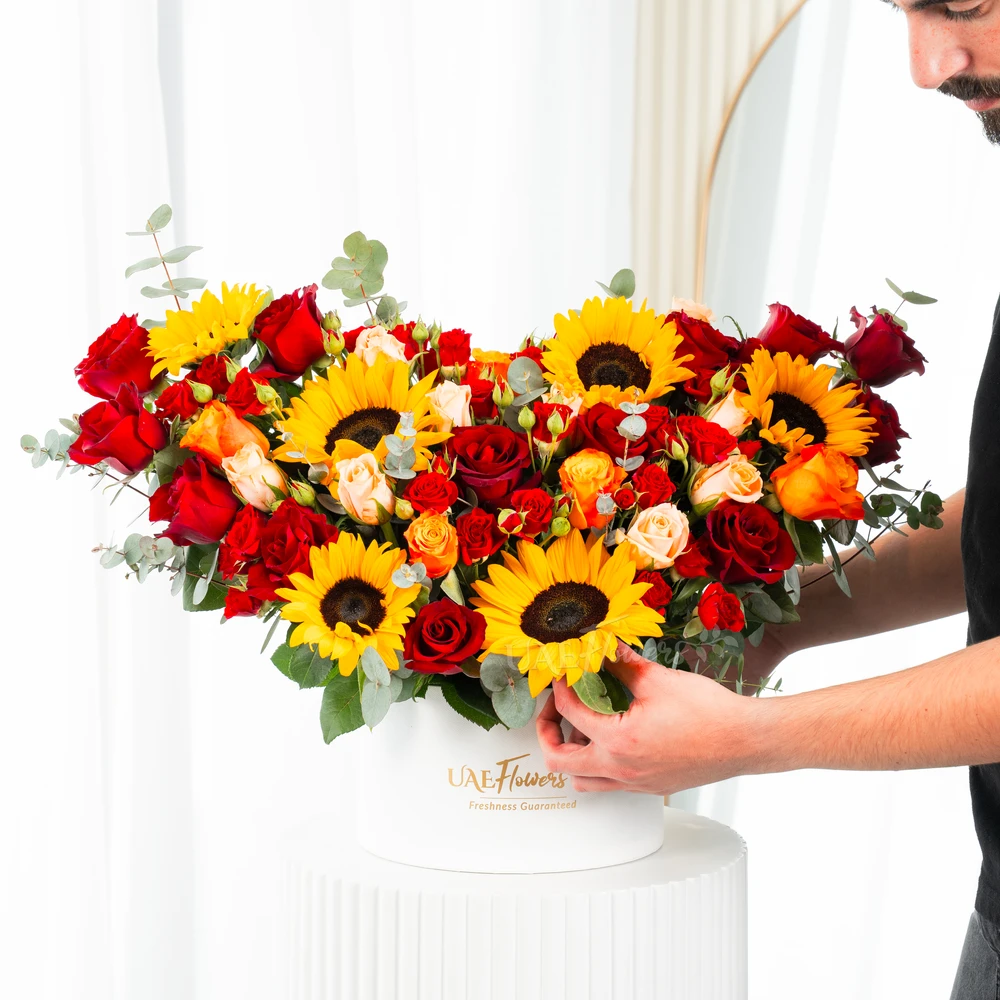 A box arrangement of 6 sunflowers, 12 red roses, 3 red spray roses, 2 orange spray roses and 2 pink spray roses with green fillers.