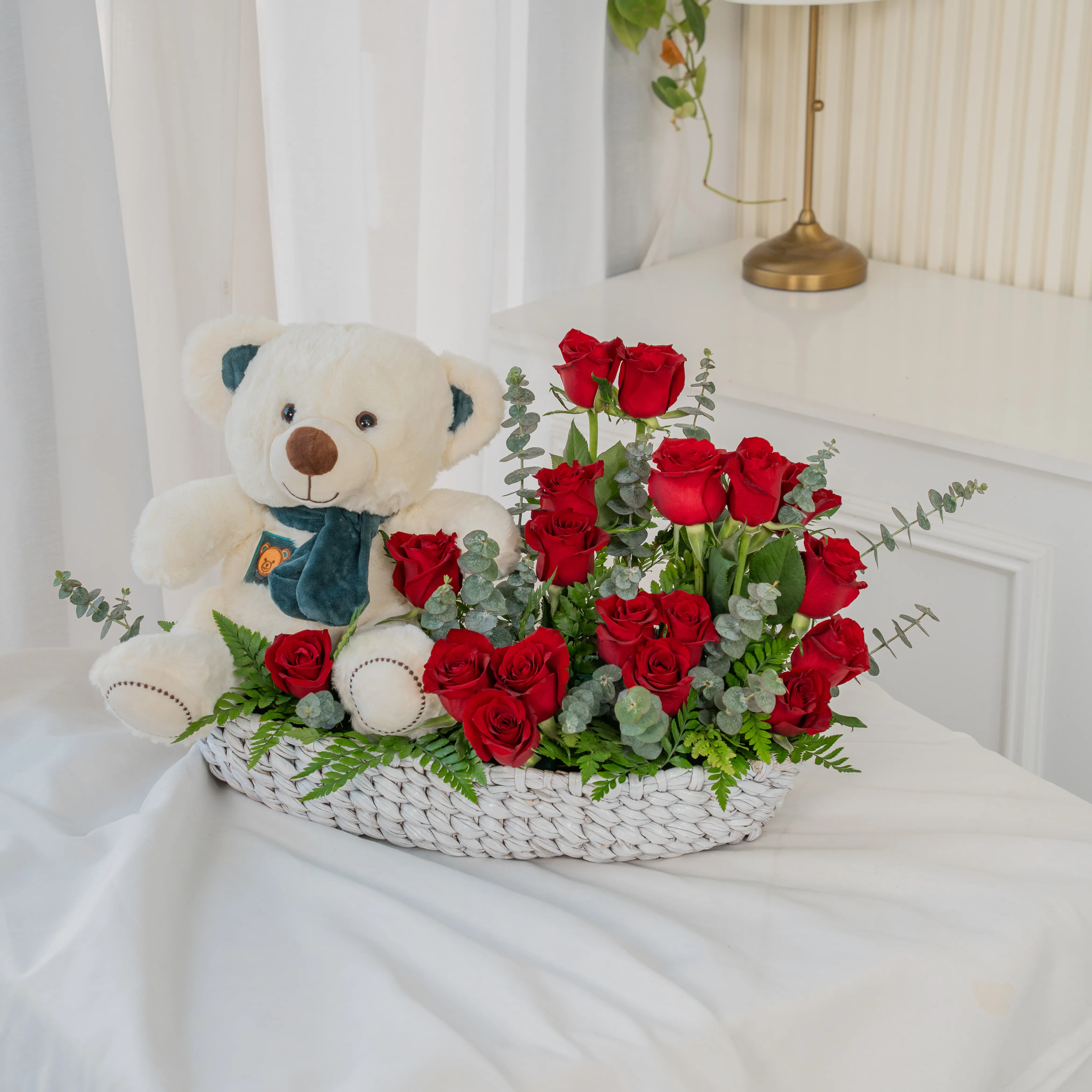 Basket arrangement of 20 red roses and small white teddy bear with eucalyptus leaf and green fillers.