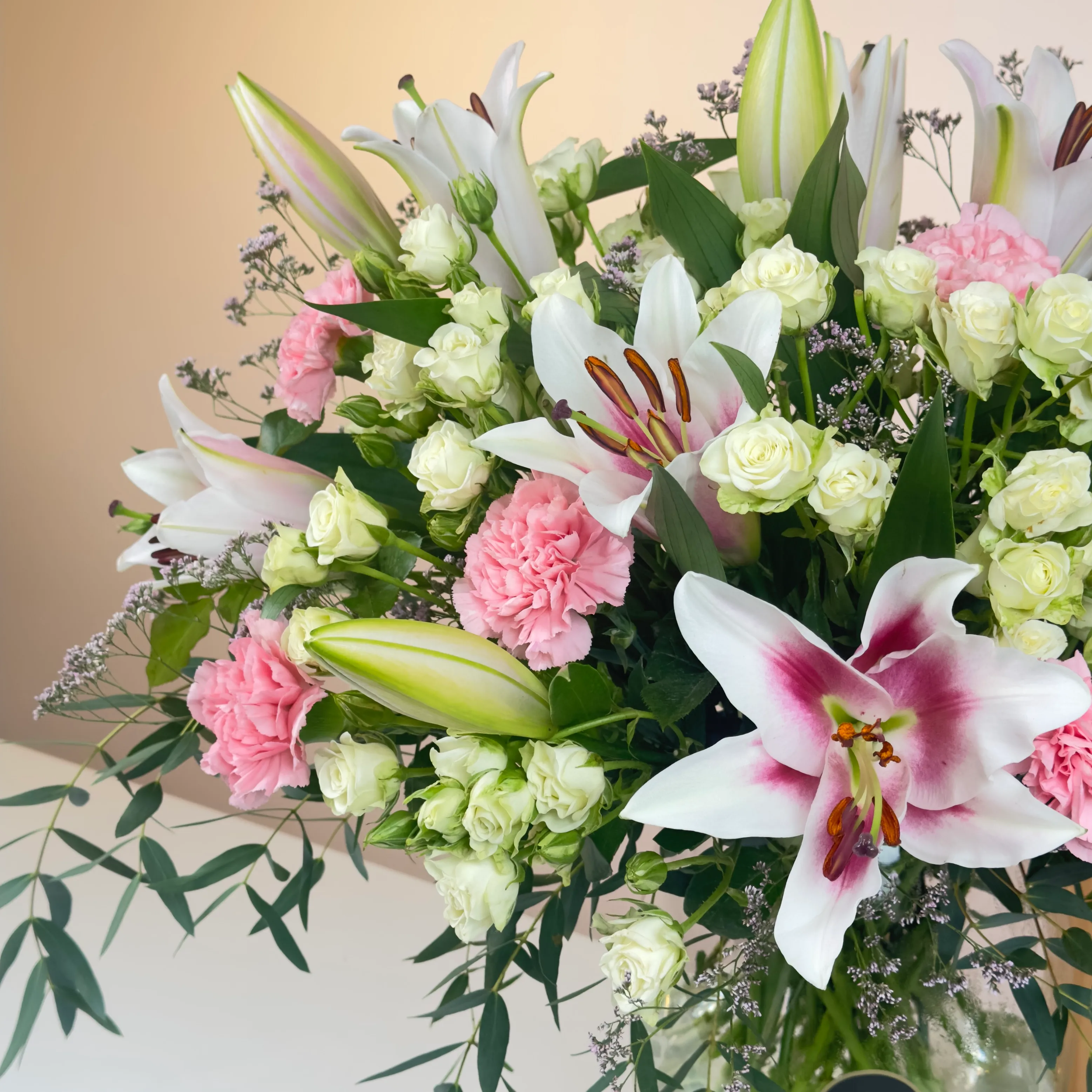 An arrangement of 4 pink lilies, 10 pink carnations, and 10 white spray roses with green fillers in a glass vase