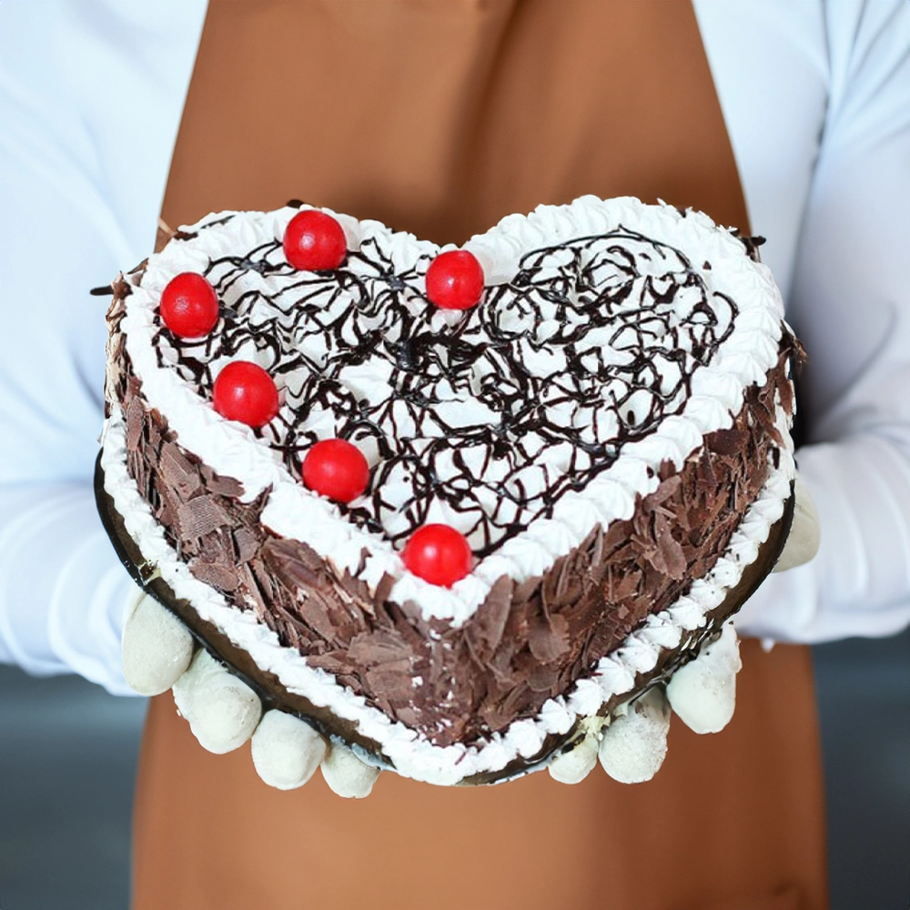 Heart shaped black forest cake with whipped cream chocolate shavings and red cherries held by a person in brown apron