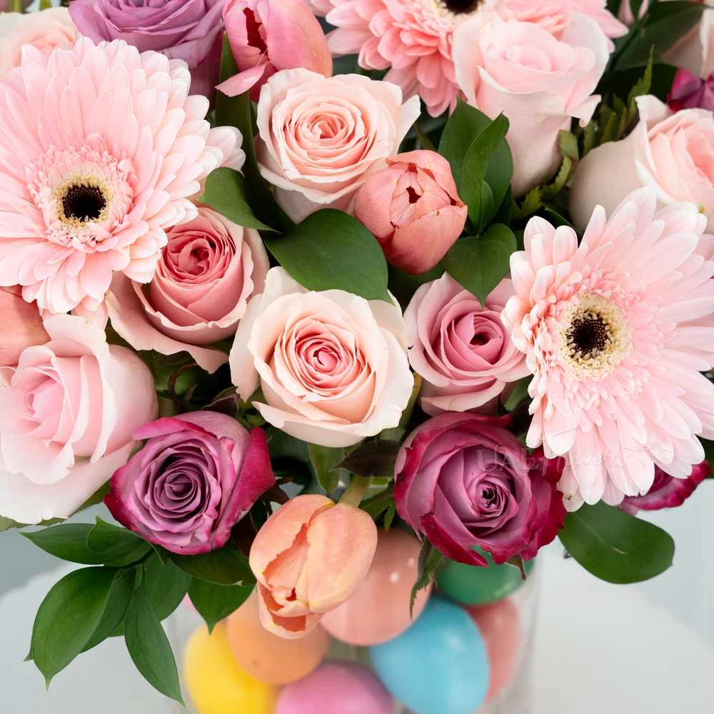 A vase arrangement of 3 pink gerberas, 7 purple roses, 15 pink roses, 10 pink tulips with green fillers and multi colored easter eggs.