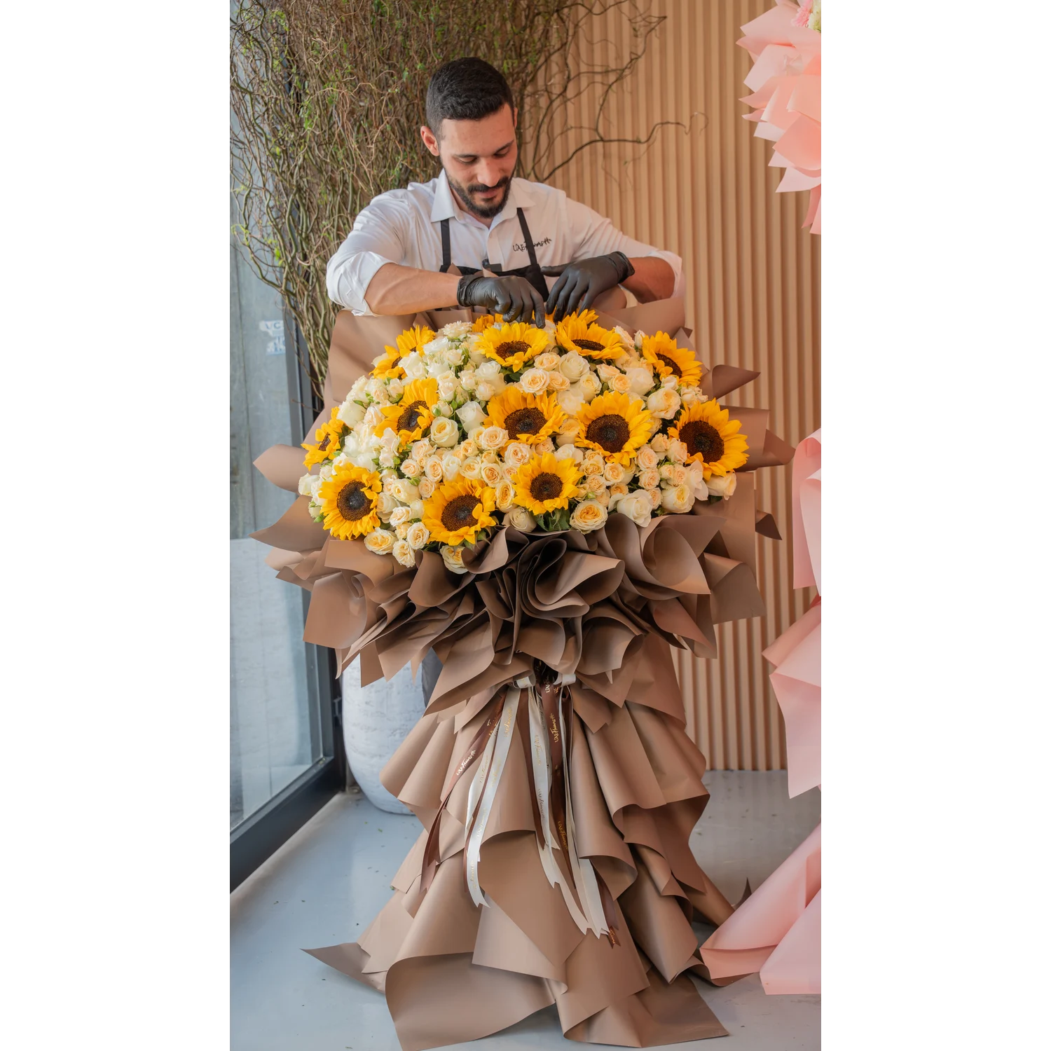 Tall luxury floral stand with 15 sunflowers, 50 spray roses, and 50 premium roses, wrapped in brown draping with satin ribbons – elegant event display by UAE Flowers.