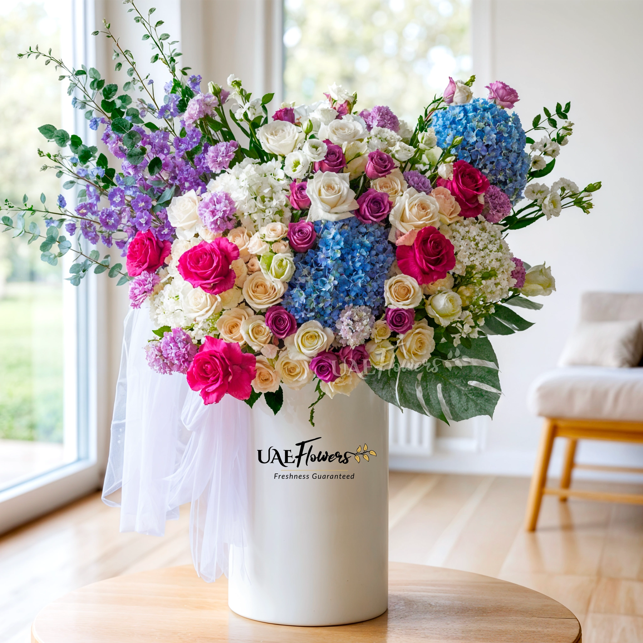 An arrangement of blue hydrangeas, white hydrangeas, purple roses, white roses, purple lisianthus, white spray roses, blue delphinium, fushia roses with green fillers and blue net in a tall round box.