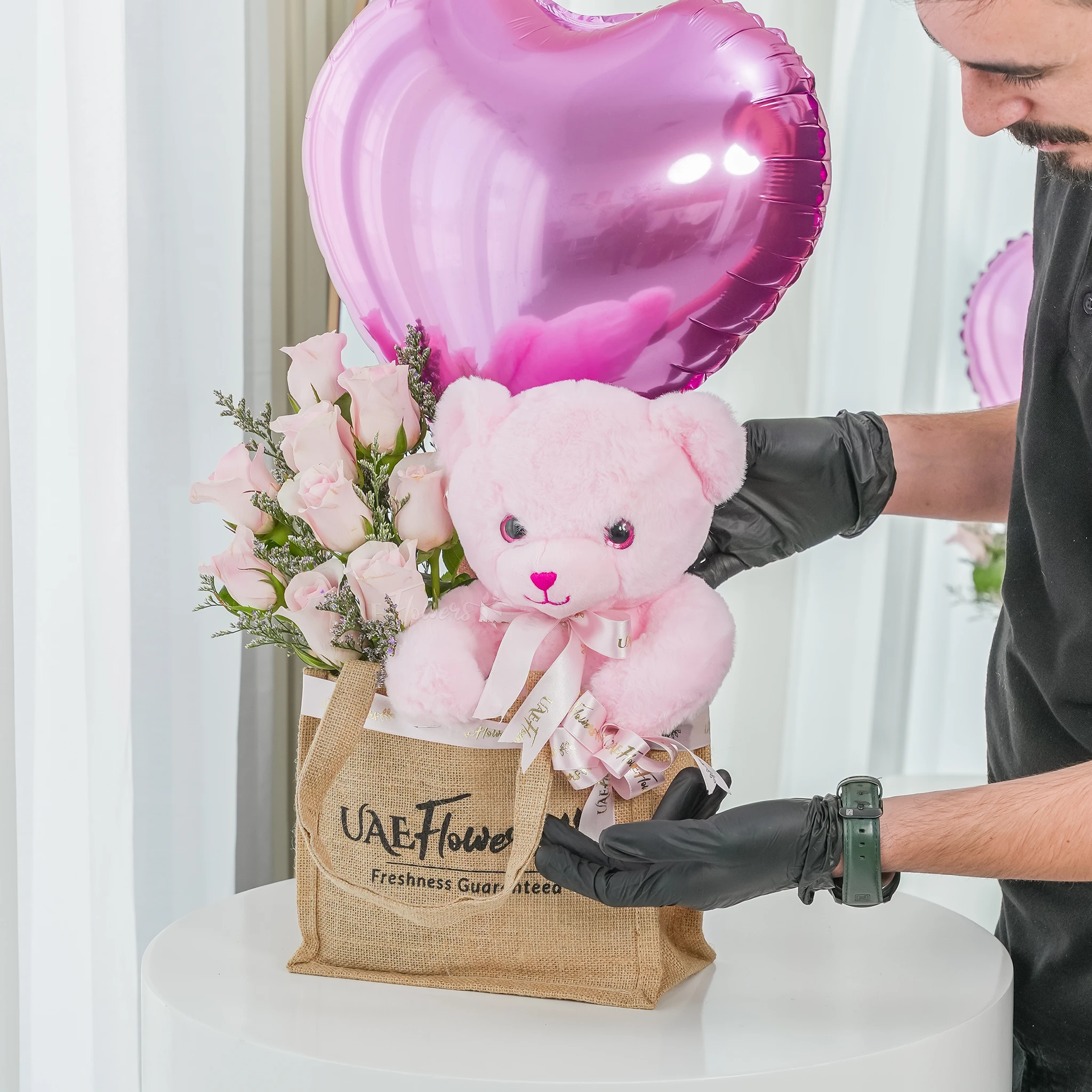 An arrangement of 7 pink roses, small size teddy bear and pink heart shaped balloons in a jute bag